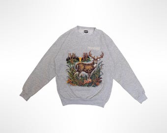 May include: A light gray crewneck sweatshirt featuring a graphic print of a deer standing in a grassy landscape with trees and a sunset in the background. The word TENNESSEE is printed in white capital letters above the deer illustration.