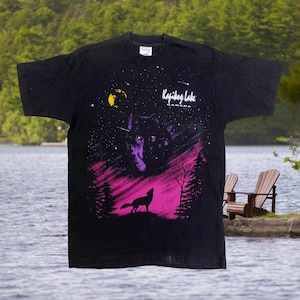 Può includere: T-shirt nera con un lupo che ulula sotto un cielo stellato e una luna gialla. Il design include un paesaggio viola e rosa con alberi. Il testo "Kajibog Lake Canada" è stampato sopra l'immagine del lupo.