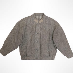 Op de afbeelding: Een grijs tweed bomberjack met een ronde halslijn, een knoopsluiting aan de voorzijde met vijf zichtbare knopen, lange mouwen en een geribbelde zoom. De binnenvoering van de kraag heeft een subtiel beige en grijs ruitpatroon.