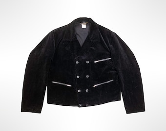 May include: A black double-breasted jacket featuring a textured finish, wide lapels, and long sleeves. The front includes two horizontal zippered pockets on the chest and waist area, along with a central button closure system with dark buttons.