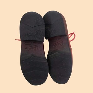May include: A pair of dark red shoes shown from the bottom view, featuring black rubber outsoles with a chevron tread pattern and a low heel. The brand name Paraboot is embossed on the center of each sole, along with the text Made in France.