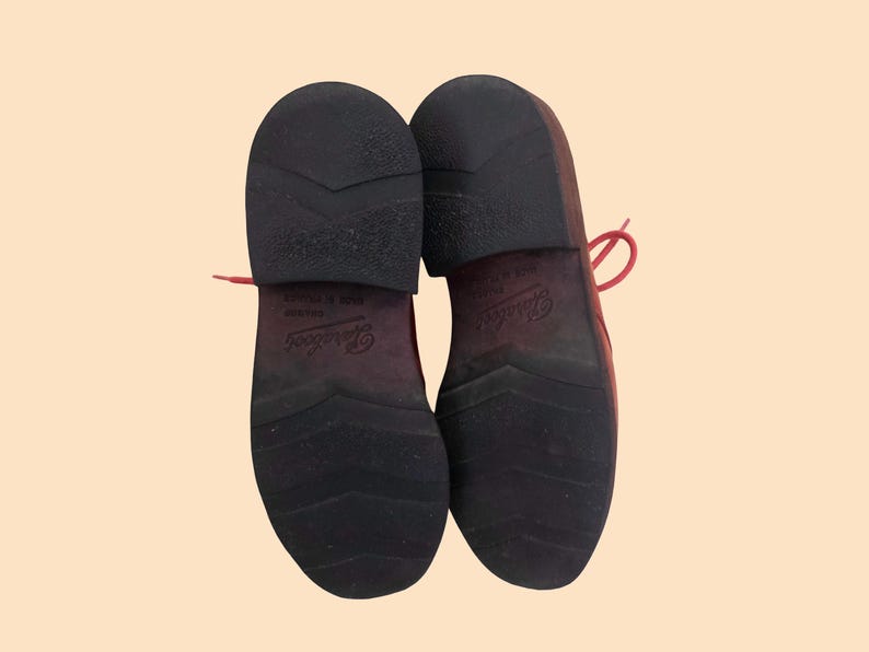 May include: A pair of dark red shoes shown from the bottom view, featuring black rubber outsoles with a chevron tread pattern and a low heel. The brand name Paraboot is embossed on the center of each sole, along with the text Made in France.