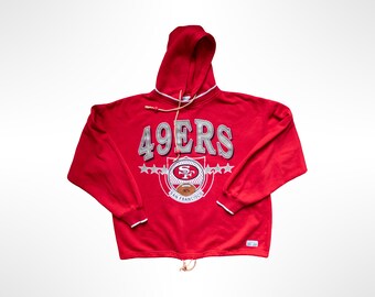 May include: A bright red hooded sweatshirt featuring the text 49ERS in silver lettering across the chest. Below the text is a circular San Francisco 49ers team logo with a football graphic, flanked by silver stars and white trim on the hood and cuffs.