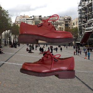 May include: A pair of red leather lace-up shoes featuring a moccasin-style toe construction, a small branded tag on the side, and a thick, dark brown rubber sole. The shoes are presented with matching red laces tied in a bow at the top.