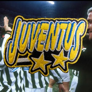 May include: Embroidered Juventus patch featuring yellow and blue lettering and two yellow stars. The patch is set against a backdrop of a football match with players in black and white striped shirts.