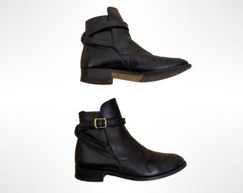 May include: A pair of black leather ankle boots featuring a pointed toe, a low block heel, and a thin strap that wraps around the ankle. The strap is secured with a small gold-toned metal buckle on the side, and the sole has a natural wood finish.
