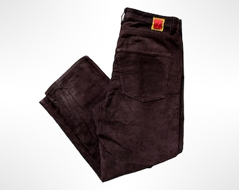 May include: Dark brown corduroy pants featuring a textured ribbed fabric, a single back pocket, and a small rectangular yellow label with red text stitched onto the waistband above the pocket. The pants are folded at an angle.