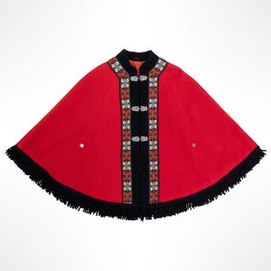 Puede incluir: Una capa de color rojo brillante con cuello negro y ribete negro a lo largo de la abertura frontal. Los paneles frontales están decorados con intrincados bordados en blanco, rojo y verde. El dobladillo tiene flecos negros y botones plateados.