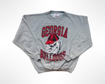 May include: A heather gray crewneck sweatshirt featuring a graphic of a bulldog wearing a red and black striped cap. The text GEORGIA BULLDOGS is printed in red with black outlines above and below the bulldog. Athens, Georgia is printed below.