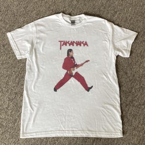 Könnte beinhalten: Ein weißes kurzärmeliges T-Shirt mit einem Grafikdruck einer Person in einem roten Anzug, die eine E-Gitarre spielt. Über der Figur ist der Text TAKANAKA in einer roten, leicht verschwommenen Schriftart aufgedruckt. Das Shirt hat einen Rundhalsausschnitt.