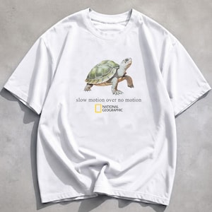 Puede incluir: Una camiseta blanca de manga corta con una ilustración gráfica de una tortuga en el centro. Debajo de la tortuga, el texto dice "slow motion over no motion" seguido del logotipo de National Geographic en su rectángulo amarillo.