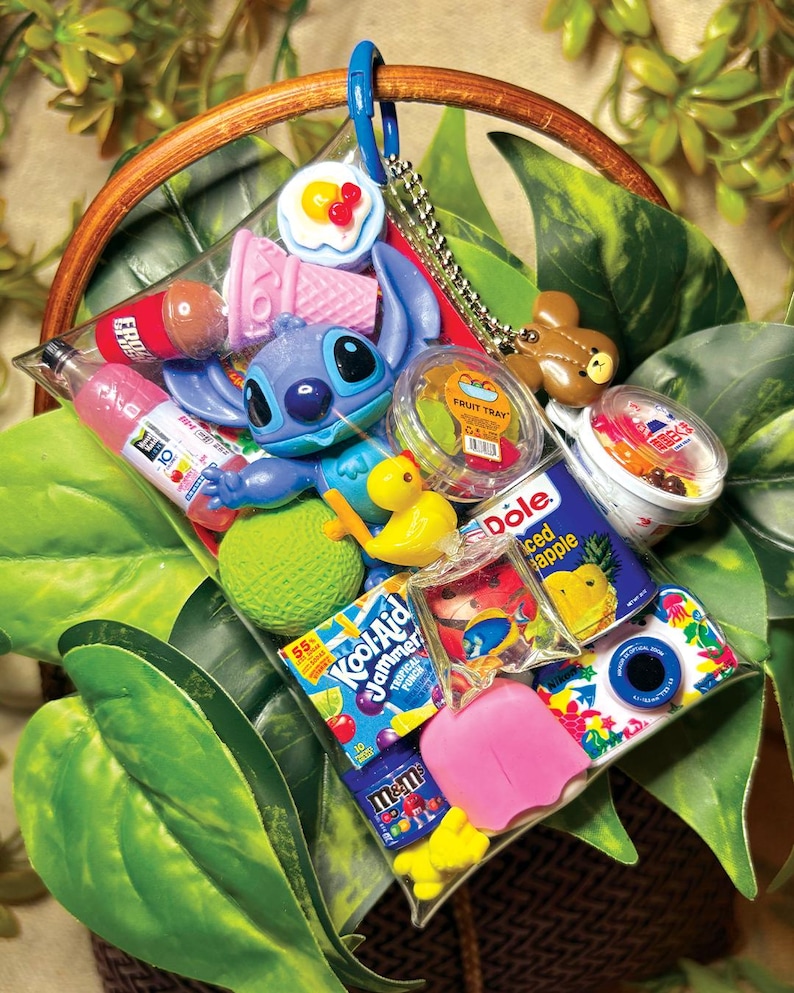K&ouml;nnte beinhalten: Ein durchsichtiger Kunststoffbeutel mit blauem Karabinerhaken, der verschiedene Miniaturgegenst&auml;nde enth&auml;lt, darunter eine blaue Stitch-Figur, eine gelbe Ente, einen Kool-Aid Jammer-Beutel, eine Dole-Ananasdose, eine kleine Kamera und eine M&M's-T&uuml;te.