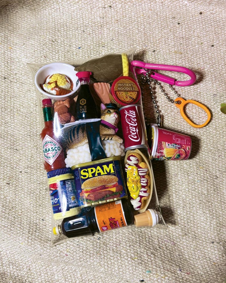 May include: A clear plastic pouch filled with miniature food-themed items including a Tabasco bottle, a Spam can, a Coca-Cola can, a bowl of noodles, a hot dog, and a cat figurine. The pouch features a pink carabiner and a silver ball chain attachment.