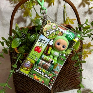 May include: A clear plastic keychain filled with miniature snacks and a green doll. The keychain includes a Juicy Juice box, Hello Panda snacks, Mike and Ike candies, and other small items. The keychain is attached to a green heart-shaped clip.