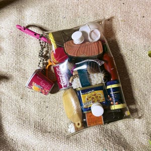 May include: A clear plastic pouch filled with miniature food-themed items, including a Coca-Cola can, a Spam tin, a sushi piece, a bottle of hot sauce, a yellow bottle, and a small cup. A pink carabiner and a silver ball chain are attached to the top.