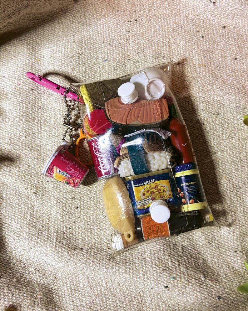 May include: A clear plastic pouch filled with miniature food-themed items, including a Coca-Cola can, a Spam tin, a sushi piece, a bottle of hot sauce, a yellow bottle, and a small cup. A pink carabiner and a silver ball chain are attached to the top.