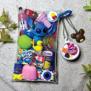 K&ouml;nnte beinhalten: Ein durchsichtiger Kunststoffbeutel gef&uuml;llt mit Miniaturspielzeug, darunter eine blaue Stitch-Figur, ein Kool-Aid Jammers-Beutel, eine Dole-Ananasdose, eine rosa Eist&uuml;te, eine gelbe Ente, ein gr&uuml;ner Ball und eine Kamera. Ein runder Schl&uuml;sselanh&auml;nger mit japanischem Text und ein brauner B&auml;renanh&auml;nger sind befestigt.