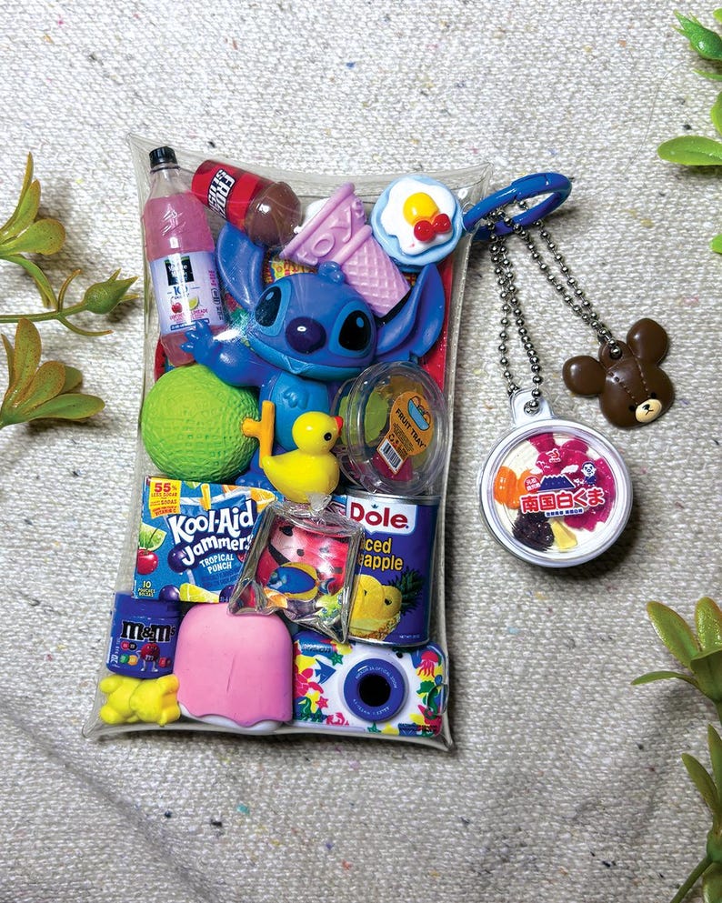 K&ouml;nnte beinhalten: Ein durchsichtiger Kunststoffbeutel gef&uuml;llt mit Miniaturspielzeug, darunter eine blaue Stitch-Figur, ein Kool-Aid Jammers-Beutel, eine Dole-Ananasdose, eine rosa Eist&uuml;te, eine gelbe Ente, ein gr&uuml;ner Ball und eine Kamera. Ein runder Schl&uuml;sselanh&auml;nger mit japanischem Text und ein brauner B&auml;renanh&auml;nger sind befestigt.
