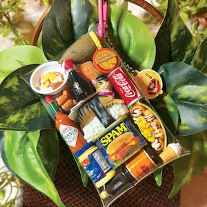 May include: A clear plastic pouch filled with miniature food-themed charms including a Coca-Cola can, a Spam tin, a Tabasco bottle, a cat figurine, instant noodles, and various other food items, all attached to a pink carabiner clip.