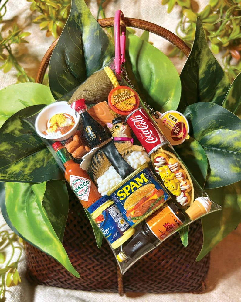 May include: A clear plastic pouch filled with miniature food-themed charms including a Coca-Cola can, a Spam tin, a Tabasco bottle, a cat figurine, instant noodles, and various other food items, all attached to a pink carabiner clip.