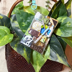Op de afbeelding: Een miniatuur sleutelhanger met een voedselthema met een verscheidenheid aan kleine items, waaronder een chocoladereep, een fles melk en een kleine cake. De sleutelhanger is bevestigd aan een gevlochten mand met groene bladeren.