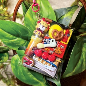 May include: A clear plastic keychain pouch filled with miniature food-themed charms including a teddy bear, a pretzel, a cup of tea, a red berry skewer, and a small box with Asian text. The pouch features a bright pink metal lobster clasp for attachment.