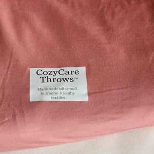 Könnte beinhalten: Nahaufnahme eines staubrosafarbenen Textils. Auf dem Stoff befindet sich ein weißes rechteckiges Etikett mit dem Text "CozyCare Throws" und dem Slogan "Made with ultra-soft, hormone-friendly textiles."