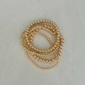 May include: Several gold beaded bracelets are displayed. The bracelets are made up of small, round beads, with variations in bead size. The gold colour is uniform across all bracelets. The bracelets are arranged in a layered style, creating a stacked effect.