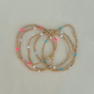 Puede incluir: Pulseras de cuentas doradas con acentos coloridos. Las pulseras presentan pequeñas cuentas doradas ensartadas, intercaladas con cuentas rosas, blancas y azules. Las pulseras están dispuestas en un estilo en capas, creando un aspecto delicado y elegante.