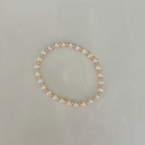 Könnte beinhalten: Ein zartes Armband mit einer Kette aus weißen, unregelmäßig geformten Perlen, die mit kleinen, goldfarbenen Perlen durchsetzt sind. Das Armband wird auf einer hellen Oberfläche präsentiert.