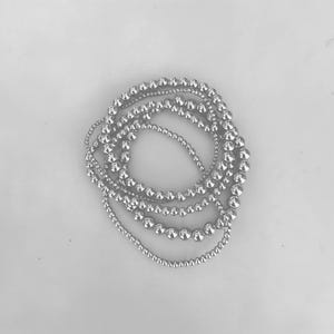 Puede incluir: Una colección de collares de cuentas plateadas de varios tamaños, dispuestos en un patrón circular. Las cuentas tienen un acabado metálico brillante, sobre un fondo blanco liso. Los collares son probablemente joyas.