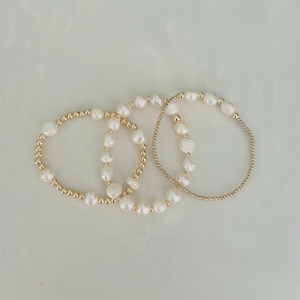 May include: Three gold beaded bracelets with white pearls. The bracelets are arranged on a white surface. The pearls vary in size and shape, adding a unique touch to each bracelet. The gold beads provide a delicate contrast to the pearls.