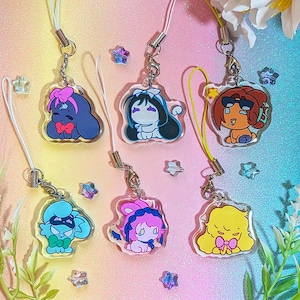 Peut inclure: Six breloques en acrylique représentant des personnages de dessins animés colorés, chacun avec un nœud et des accessoires. Chaque breloque est attachée à une lanière avec un fermoir en métal. Le fond est un dégradé de rose, de bleu et de jaune, avec des étoiles.