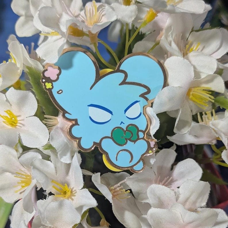 May include: A light blue enamel pin featuring a cartoon mouse design with a gold outline. The mouse has closed eyes, a green bow, and pink accents. The pin is set against a backdrop of white flowers.