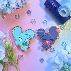 May include: Two enamel pins featuring cartoon mouse designs in light blue and dark blue, each with gold accents. The light blue mouse has a green bow, while the dark blue mouse has a pink bow. Scattered around are star-shaped beads and blue thread spools.