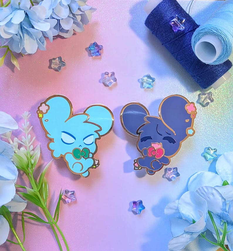 May include: Two enamel pins featuring cartoon mouse designs in light blue and dark blue, each with gold accents. The light blue mouse has a green bow, while the dark blue mouse has a pink bow. Scattered around are star-shaped beads and blue thread spools.