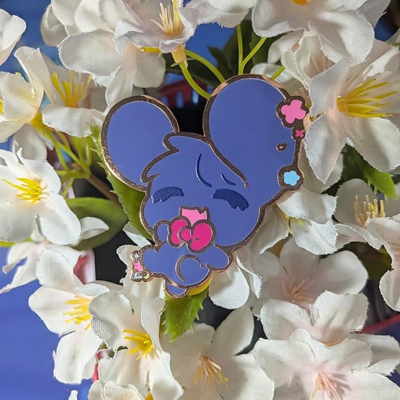 May include: A blue mouse enamel pin with gold trim, featuring a pink bow and flower accents. The pin is set against a backdrop of white flowers with yellow centers, creating a whimsical and charming aesthetic.
