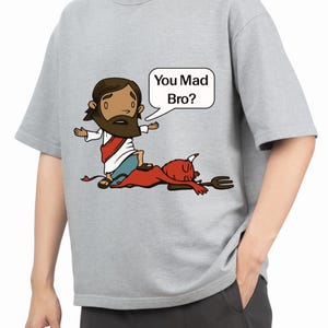 Könnte beinhalten: Hellgraues T-Shirt mit einer Cartoon-Illustration. Die Grafik zeigt eine Figur in weißer Robe, die über einem roten Teufel mit einer Mistgabel steht. Eine Sprechblase sagt: "You Mad Bro?"