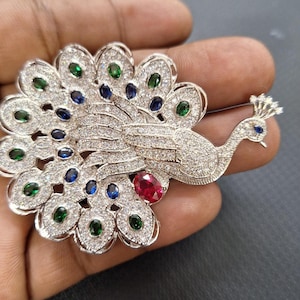 Op de afbeelding: Zilverkleurige pauwbroche, bezet met heldere en gekleurde edelstenen. De staartveren van de pauw zijn versierd met groene en blauwe stenen, terwijl een rode steen het lichaam accentueert. De broche is gedetailleerd ontworpen.