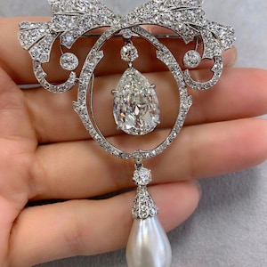 May include: A diamond and pearl brooch. The brooch features a large pear-shaped diamond pendant suspended from a diamond-encrusted oval frame. The frame is adorned with a diamond bow and additional diamond accents. A large, white pearl dangles from the bottom.