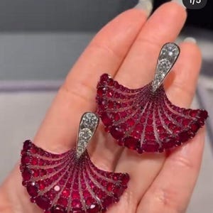 May include: A pair of ruby and diamond earrings. The earrings feature a fan-shaped design with numerous small, round rubies in a deep red hue. Each earring has a silver-colored top section with clear diamonds.