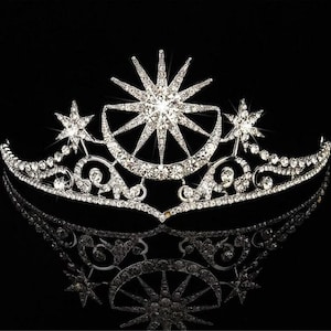 May include: A silver-toned tiara with a celestial design, featuring a large starburst, crescent moons, and smaller stars. The crown is encrusted with numerous clear crystals, reflecting light against a black background.
