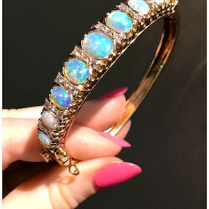 Könnte beinhalten: Ein goldener Armreif mit ovalen Opalen und kleinen Diamanten. Die Opale zeigen ein lebhaftes Farbspiel mit Blau- und Grüntönen. Das Armband ist vor einem dunklen Hintergrund platziert, was das Funkeln des Schmucks hervorhebt.