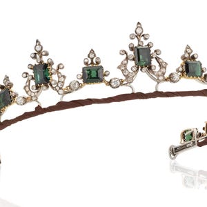 Puede incluir: Una tiara ornamentada con piedras preciosas verdes talla esmeralda y pequeños diamantes engastados en un marco de metal plateado y dorado. La tiara tiene una banda marrón y se muestra sobre un fondo blanco.