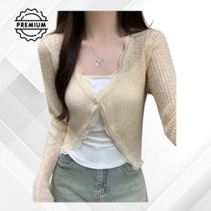 Puede incluir: Un cárdigan color crema de manga larga con un ribete de encaje festoneado. El cárdigan está abotonado en el pecho y se lleva sobre una camiseta blanca. La imagen también incluye un collar plateado y pantalones cortos de mezclilla. La palabra "PREMIUM" está en la esquina superior izquierda.