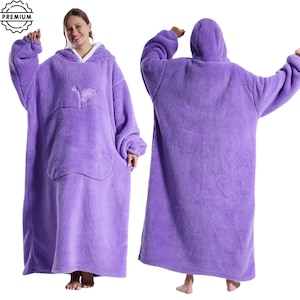 May include: A plush, oversized purple wearable blanket with a hood and a large front pocket. The blanket features a white fleece lining around the hood and a stylized apple design on the front. The blanket is designed for comfort and warmth.