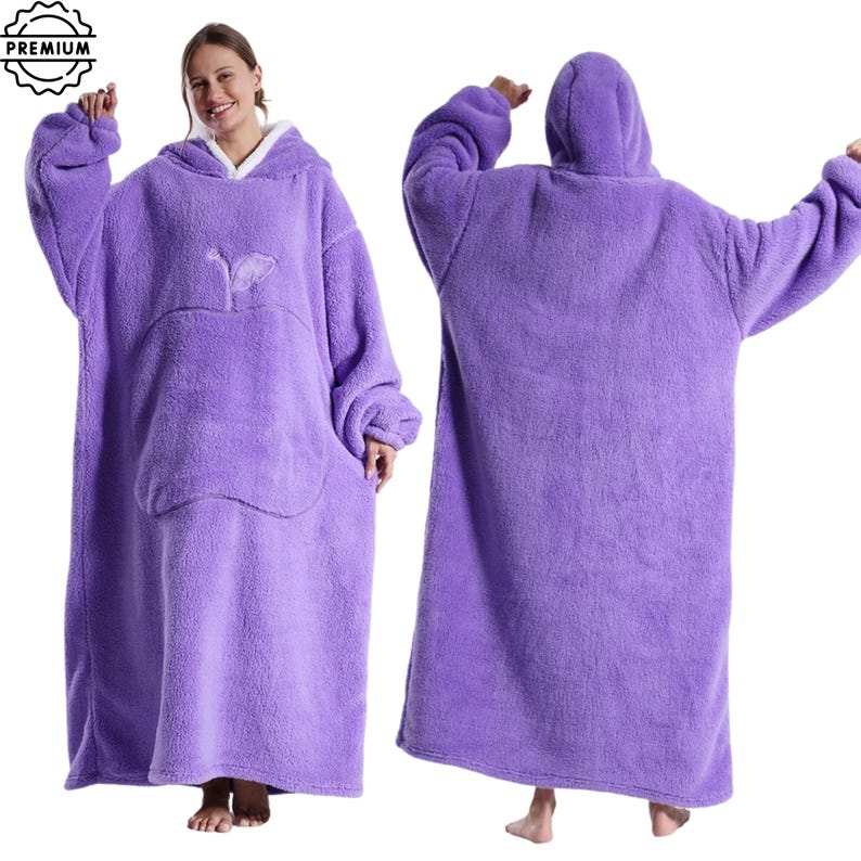 May include: A plush, oversized purple wearable blanket with a hood and a large front pocket. The blanket features a white fleece lining around the hood and a stylized apple design on the front. The blanket is designed for comfort and warmth.