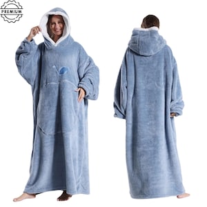 May include: A light blue, oversized wearable blanket with a hood and a large front pocket. The blanket has a soft, plush texture and a white lining in the hood. A small blue leaf design is on the front. The blanket is designed to be worn.