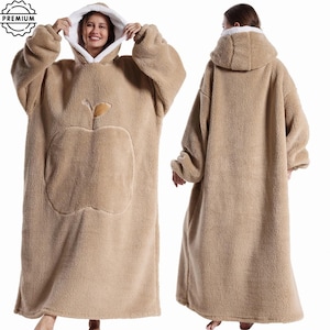 May include: A tan, oversized wearable blanket with a hood and a large apple design on the front. The blanket has a white fleece lining in the hood and is made of a soft, plush material. The image shows the blanket from the front and back.