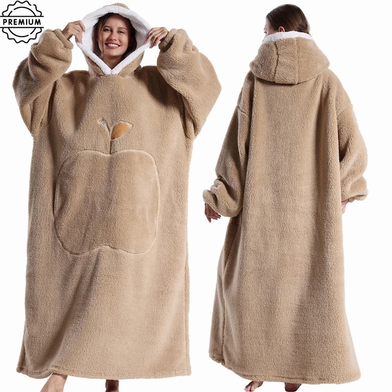 May include: A tan, oversized wearable blanket with a hood and a large apple design on the front. The blanket has a white fleece lining in the hood and is made of a soft, plush material. The image shows the blanket from the front and back.